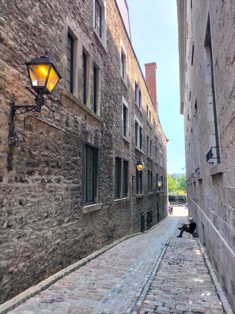 An alley on Rue Saint Paul. In the foreground, a street lamp is lit and a man in the background sitting on a chair.