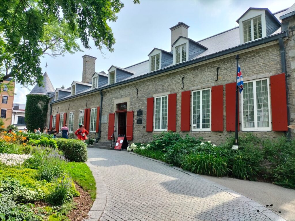 A historic stone building with red windows and a dark roof. A flowerbed in the foreground on the left.