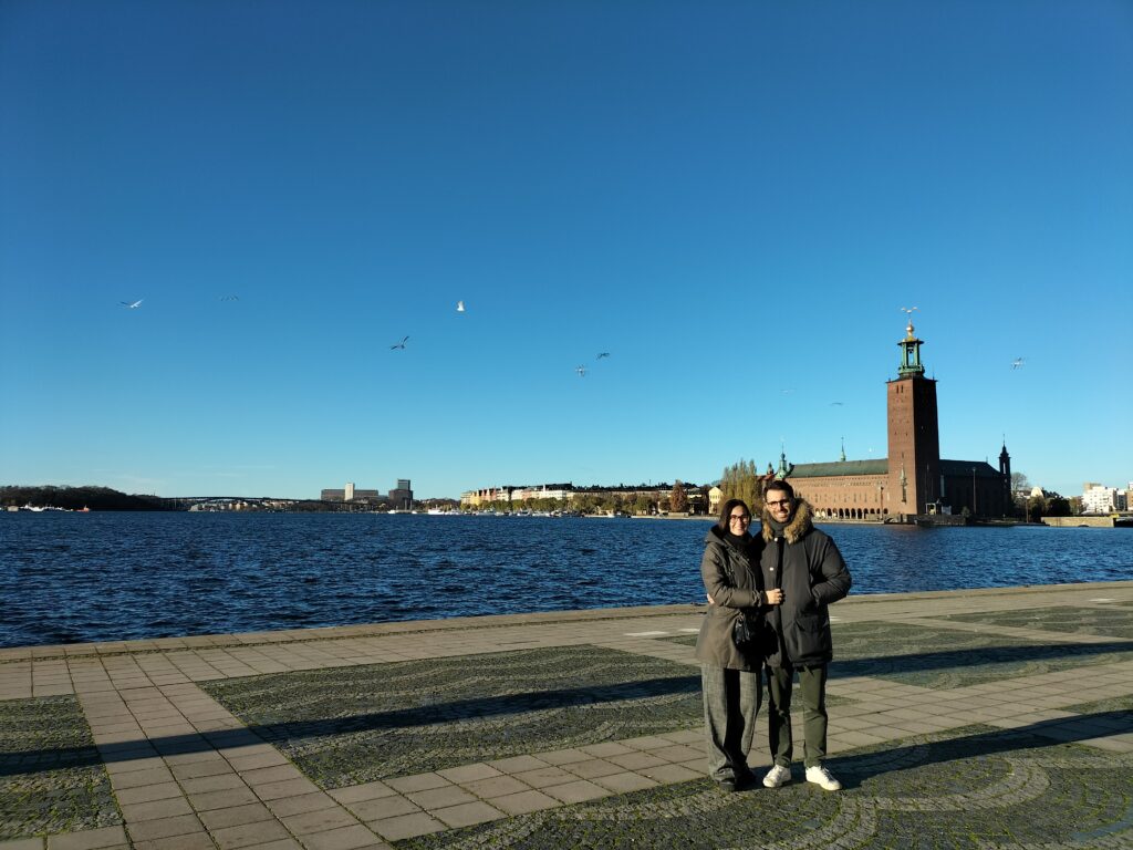 Tiziano and Claudia smiling in Stockholm's City Centre. On the background the City Hall and a blue sky.