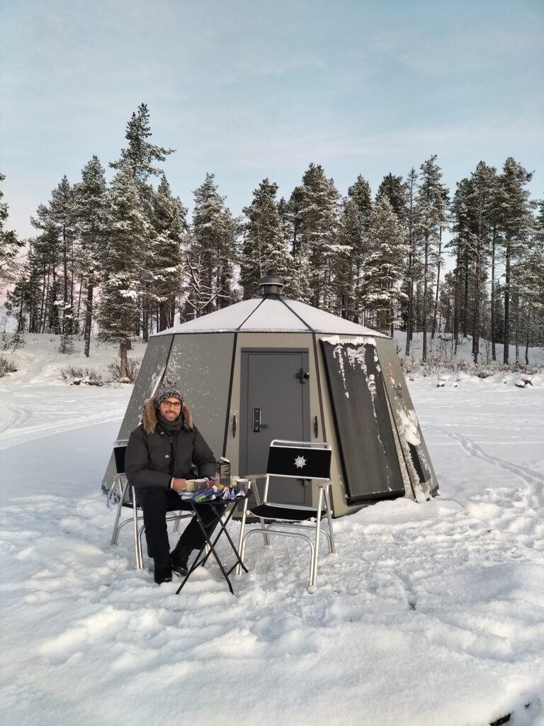 Tiziano having breakfast outside the glass igloo on the camping table. On the background the trees of the swedish forest.