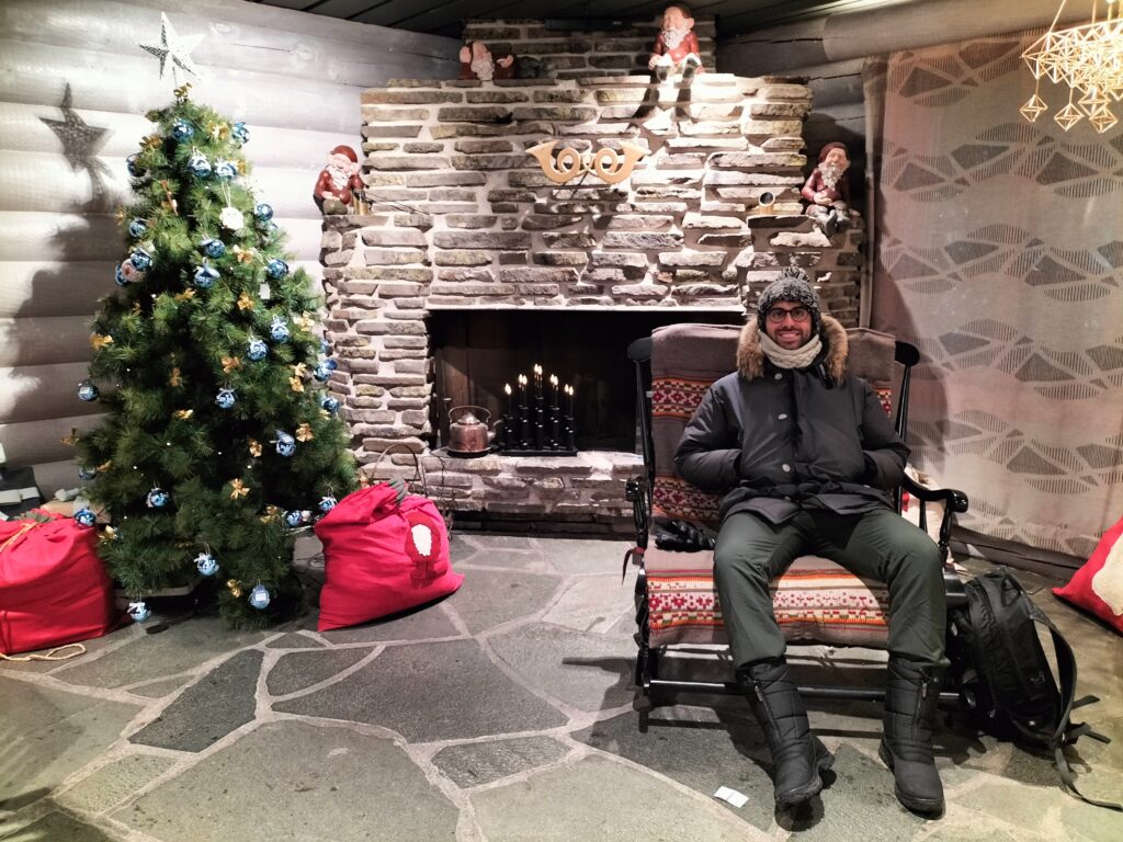 Tiziano sitting inside Santa Claus' house on a rocking chair. In the background the stone fireplace and on the left a Christmas tree with two red bags for gifts.