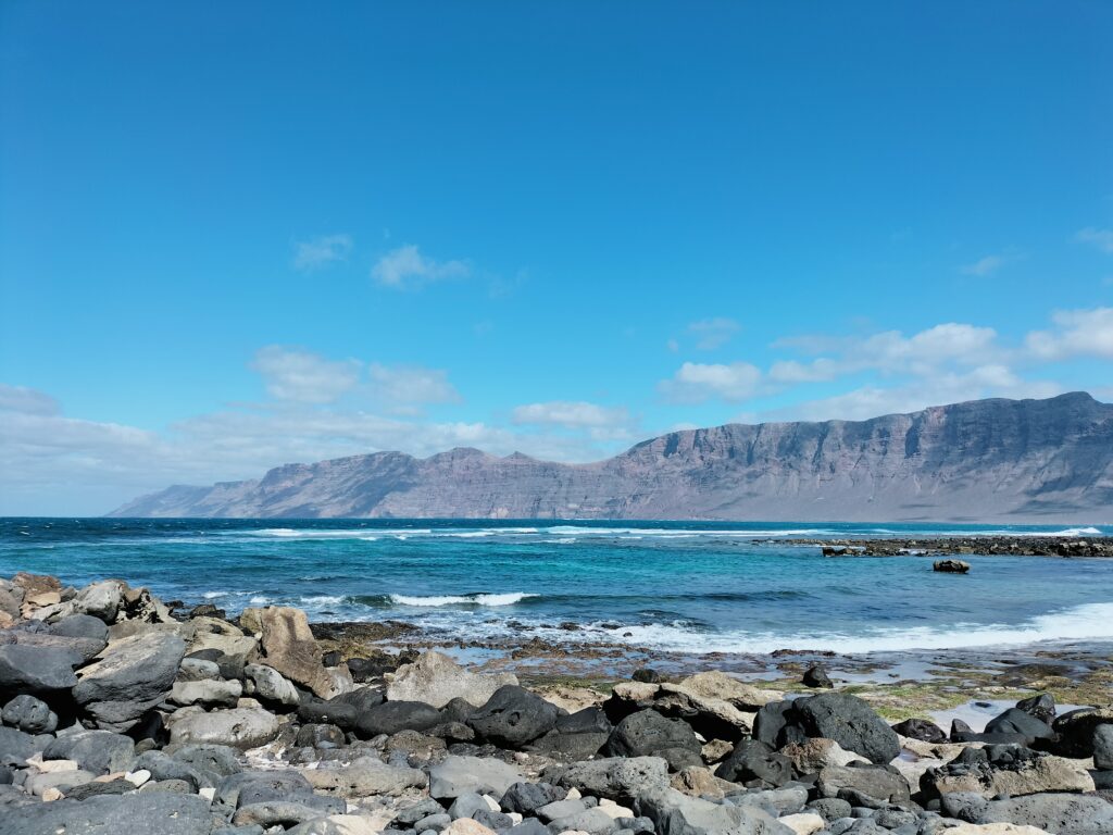 Image of the sea of ​​Famara Beach. In the foreground the stones and in the background the mountains of the island.