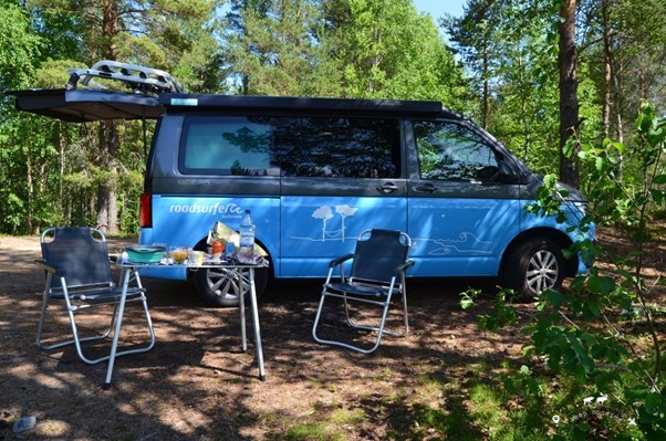 Our van parked in a spot on the lake shore in a forest a few km from the main road. A small table with two chairs in the foreground for lunch. The rear door of the van is open.