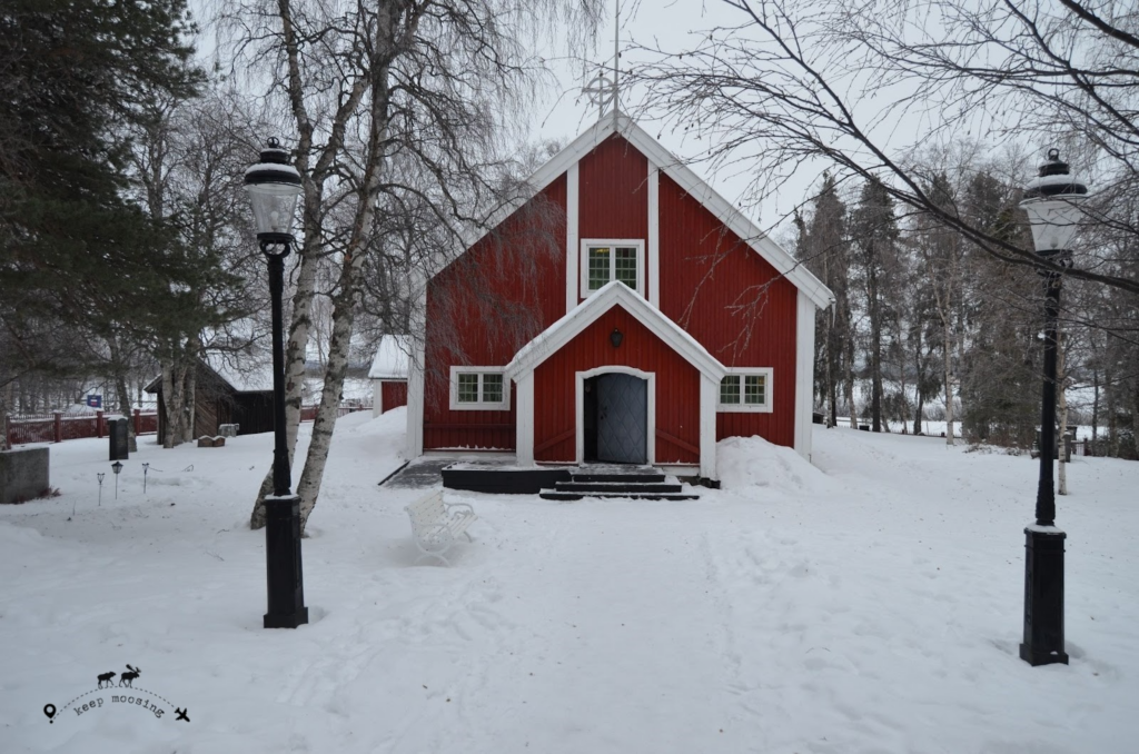 Typical red wooden church immersed in a snowy backdrop. Two snow-covered street lamps stand in front of the church entrance.