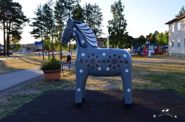 The large wooden horse typical of the Dalarna region. In the background the village of Rattvik with a house on the right and trees on the left and in the center.