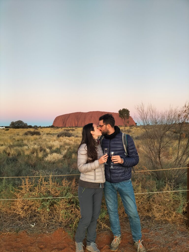 Tiziano and Claudia kissing with 2 glasses of wine standing in front of Uluru.