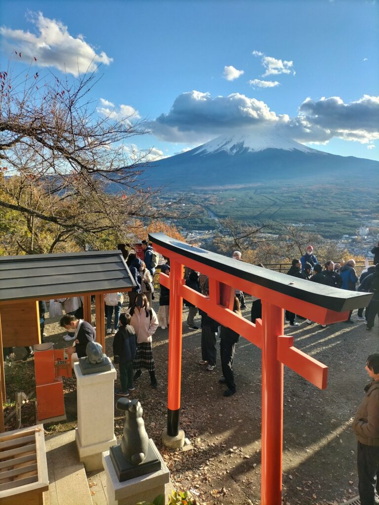 In the foreground a red torii with many tourists and in the background Mt. Fuji with clouds.